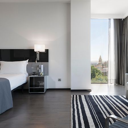 Eurostars Palace Кордоба Екстериор снимка The photo shows a modern hotel room featuring a large bed with white linens and dark cushions, paired with a sleek nightstand. There is a contemporary chair and a small table nearby, along with a striped area rug on the floor. A large window allows n