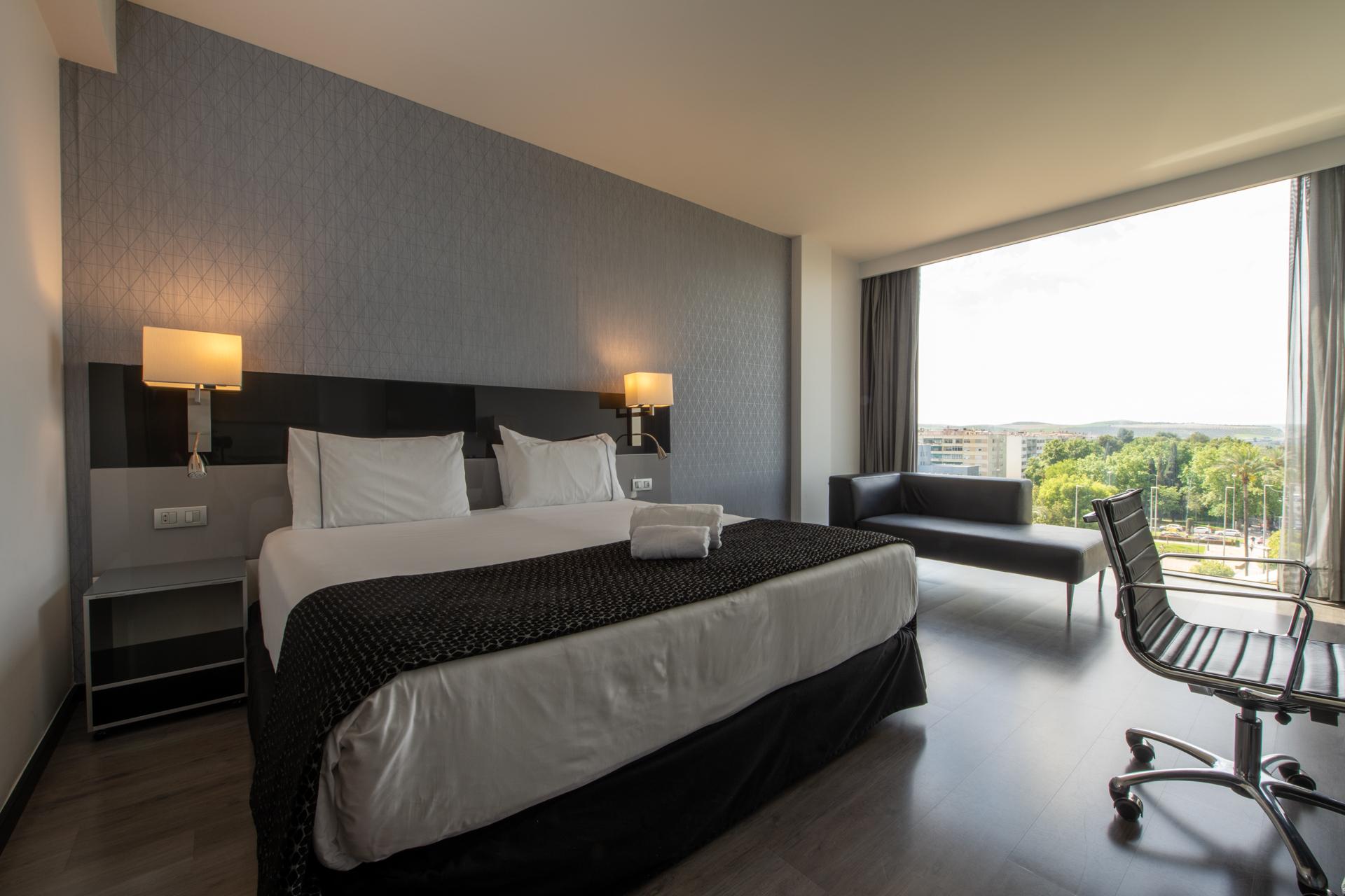 Eurostars Palace Кордоба Екстериор снимка The photo shows a modern hotel room featuring a large bed with white linens and a dark patterned throw. On either side of the bed, there are stylish wall-mounted lamps. A contemporary desk chair is positioned at a small desk, and there’s a sofa near 
