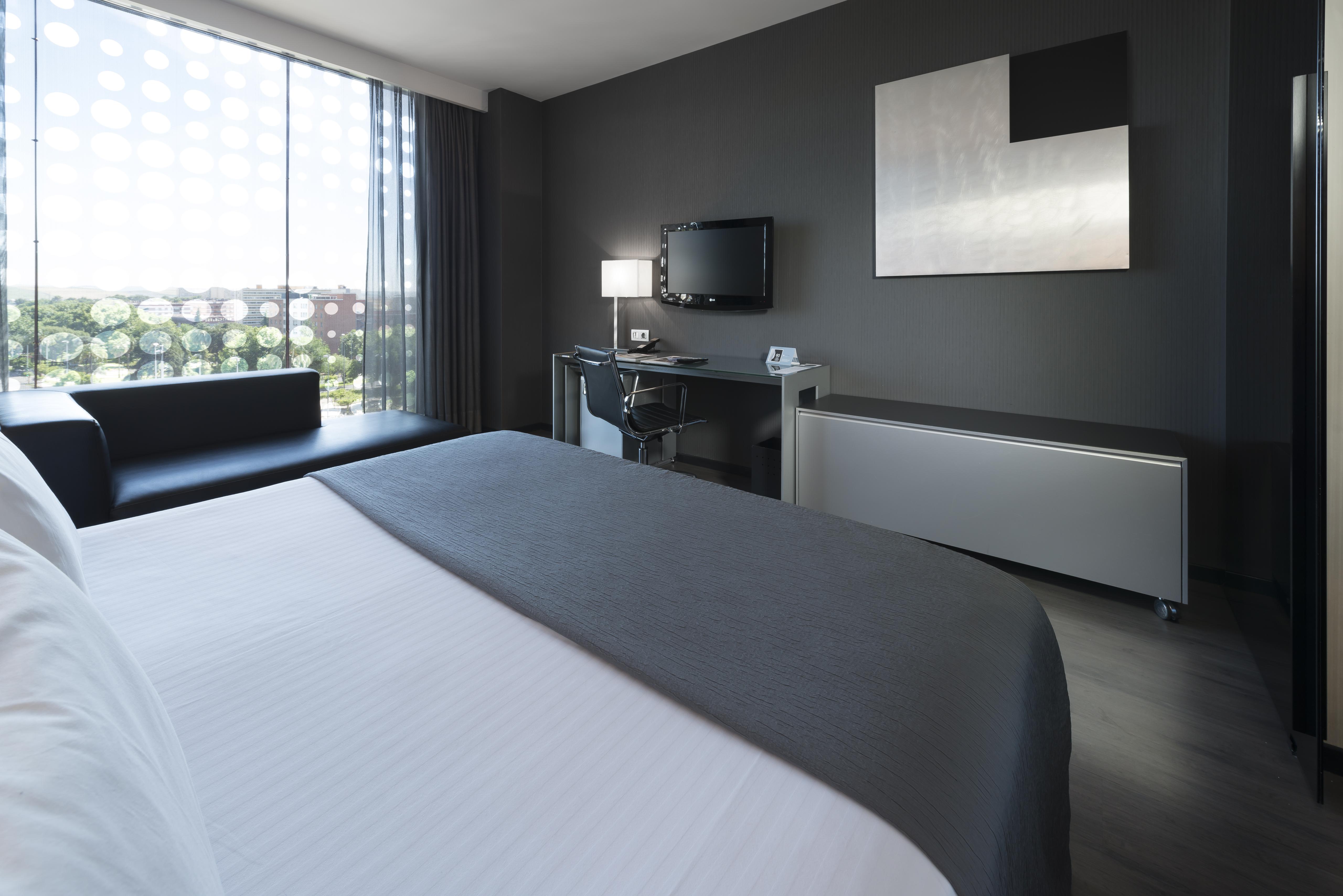 Eurostars Palace Кордоба Екстериор снимка The photo shows a modern hotel room featuring a sleek design. In the foreground, there is a bed with a white coverlet and gray accents. To the side, a dark sofa is visible. The room has a large window with sheer curtains, allowing natural light to en