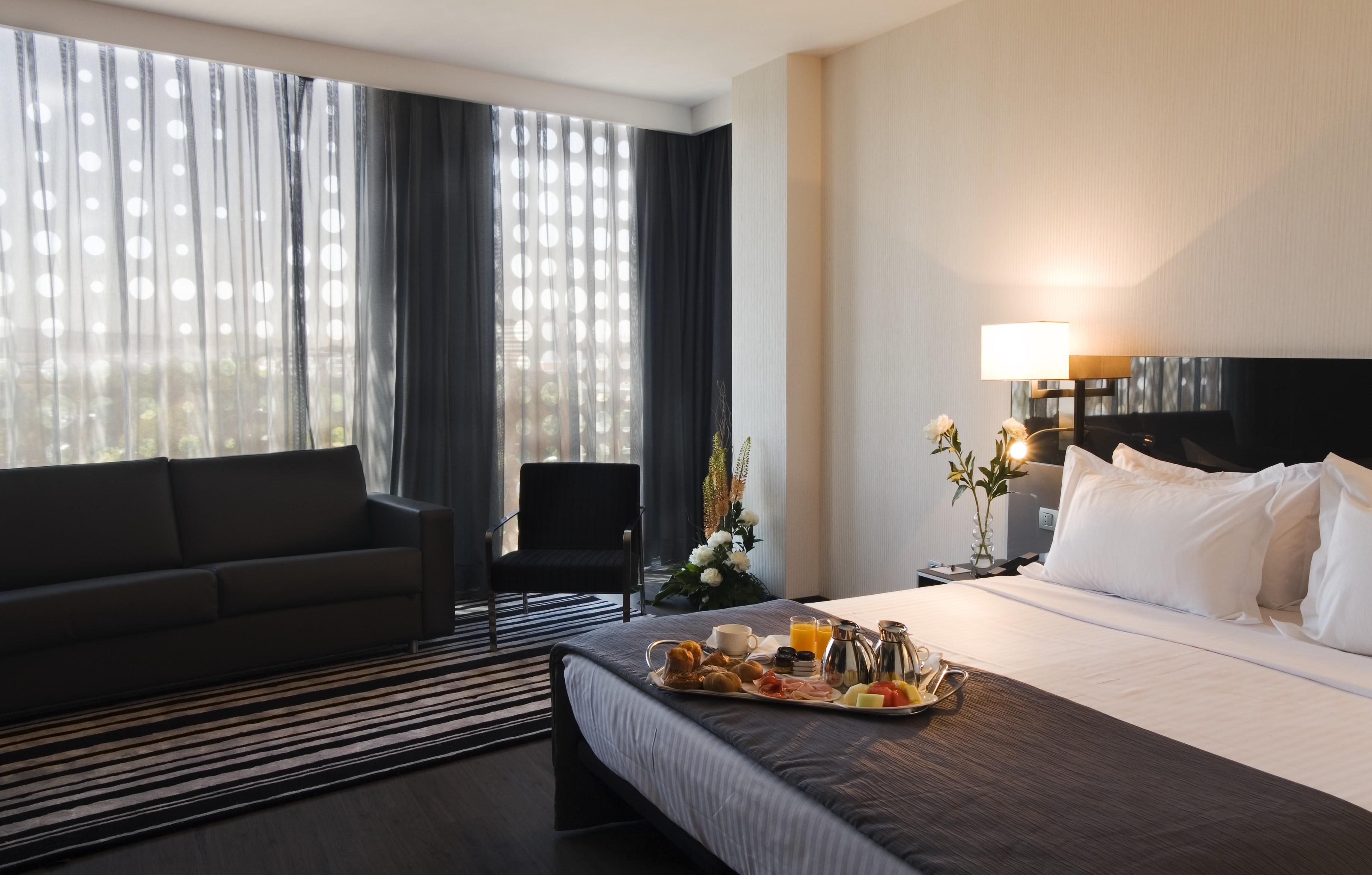 Eurostars Palace Кордоба Екстериор снимка The photo shows a modern hotel room featuring a neatly made bed with white linens and a dark grey blanket. There is a tray on the bed with a breakfast spread, including pastries, fruit, and beverages. To the left, there is a comfortable-looking couch