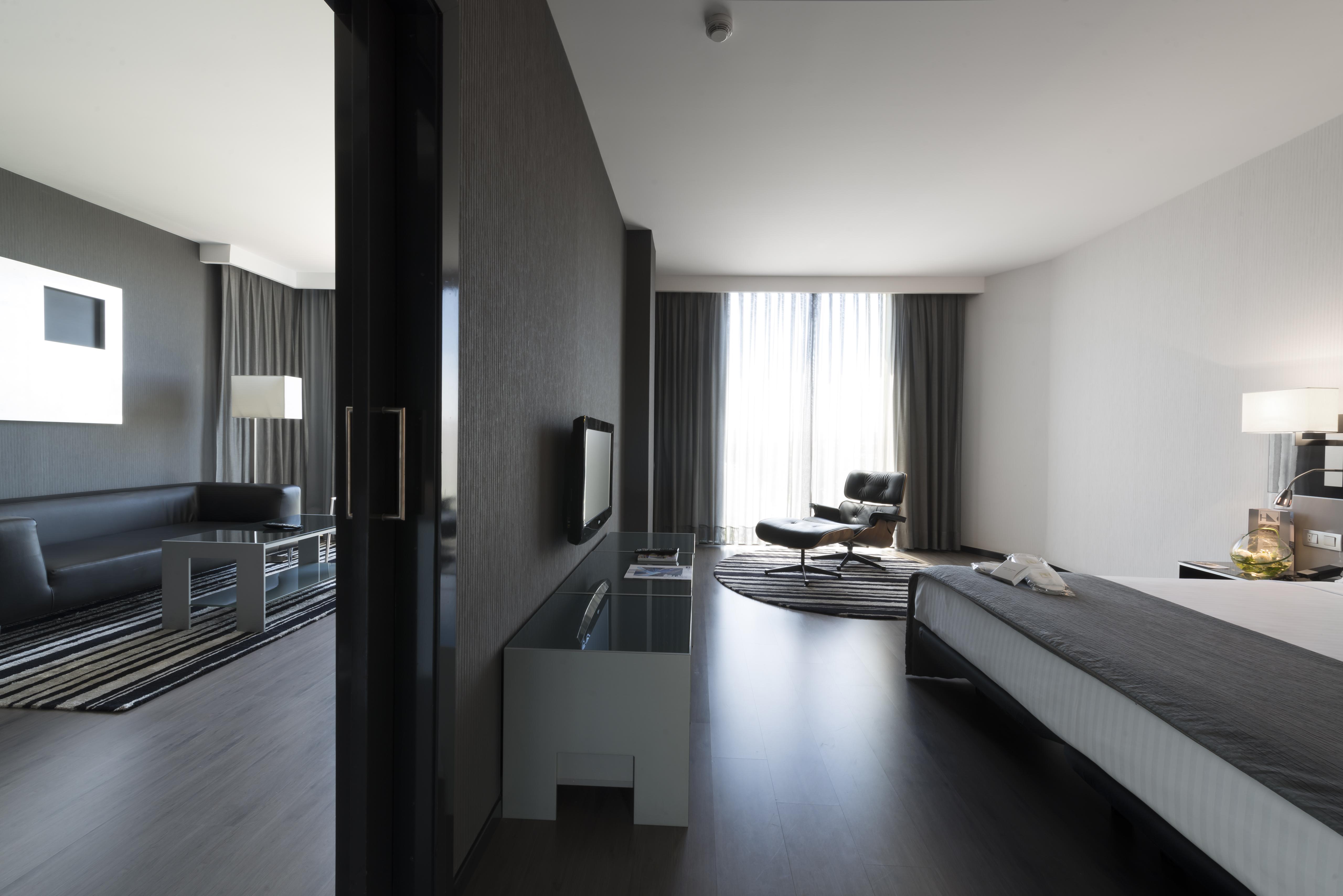 Eurostars Palace Кордоба Екстериор снимка The photo shows a modern hotel room or suite featuring a contemporary design. On the left side, there is a living area with a black sofa, a coffee table, and light fixtures. The large windows, covered with sheer curtains, let in natural light, enhanc
