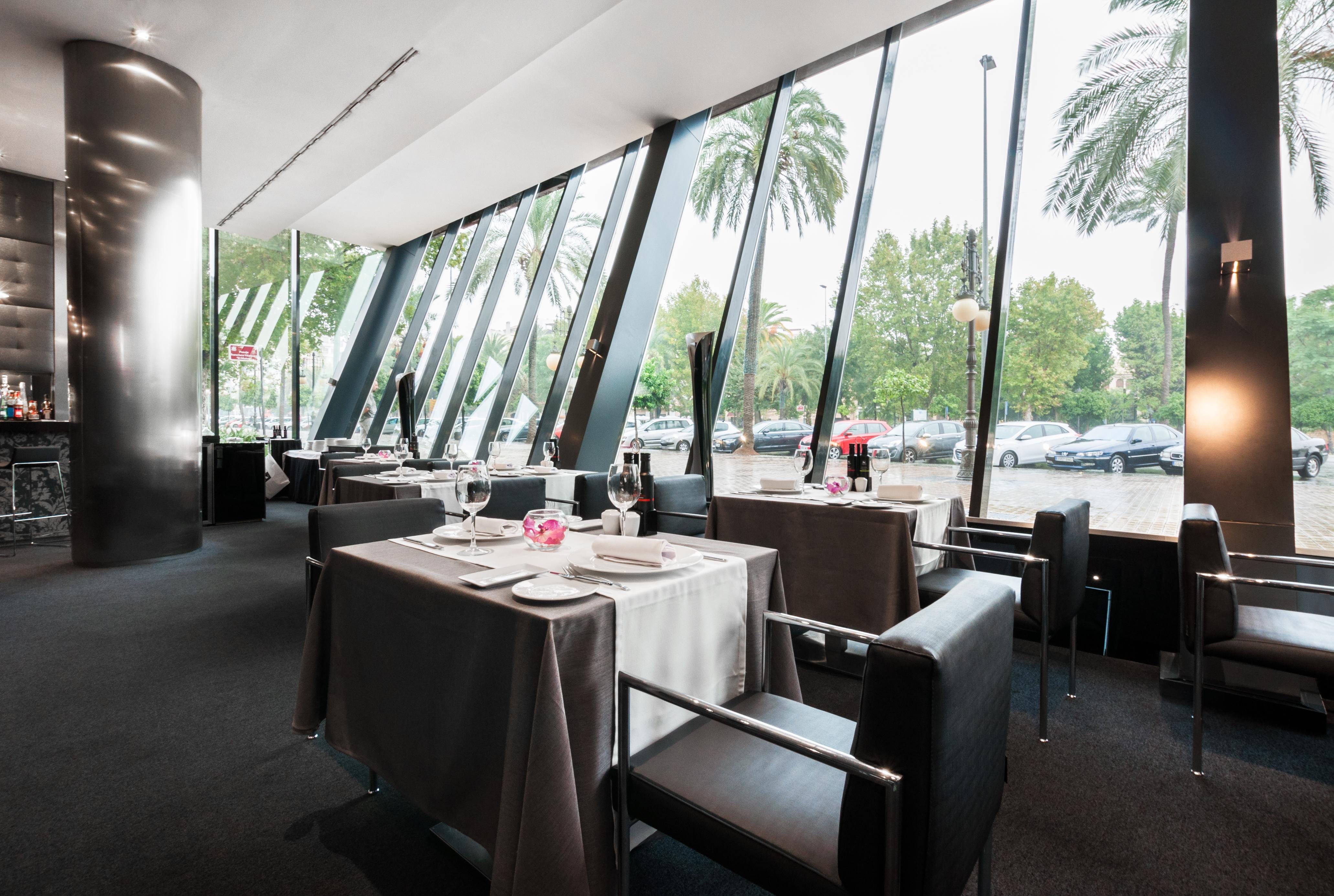 Eurostars Palace Кордоба Екстериор снимка The photo shows the interior of a modern restaurant featuring large glass windows that provide an expansive view of the outdoors, including palm trees. The dining area is elegantly set up with tables covered in gray tablecloths, decorated with simple