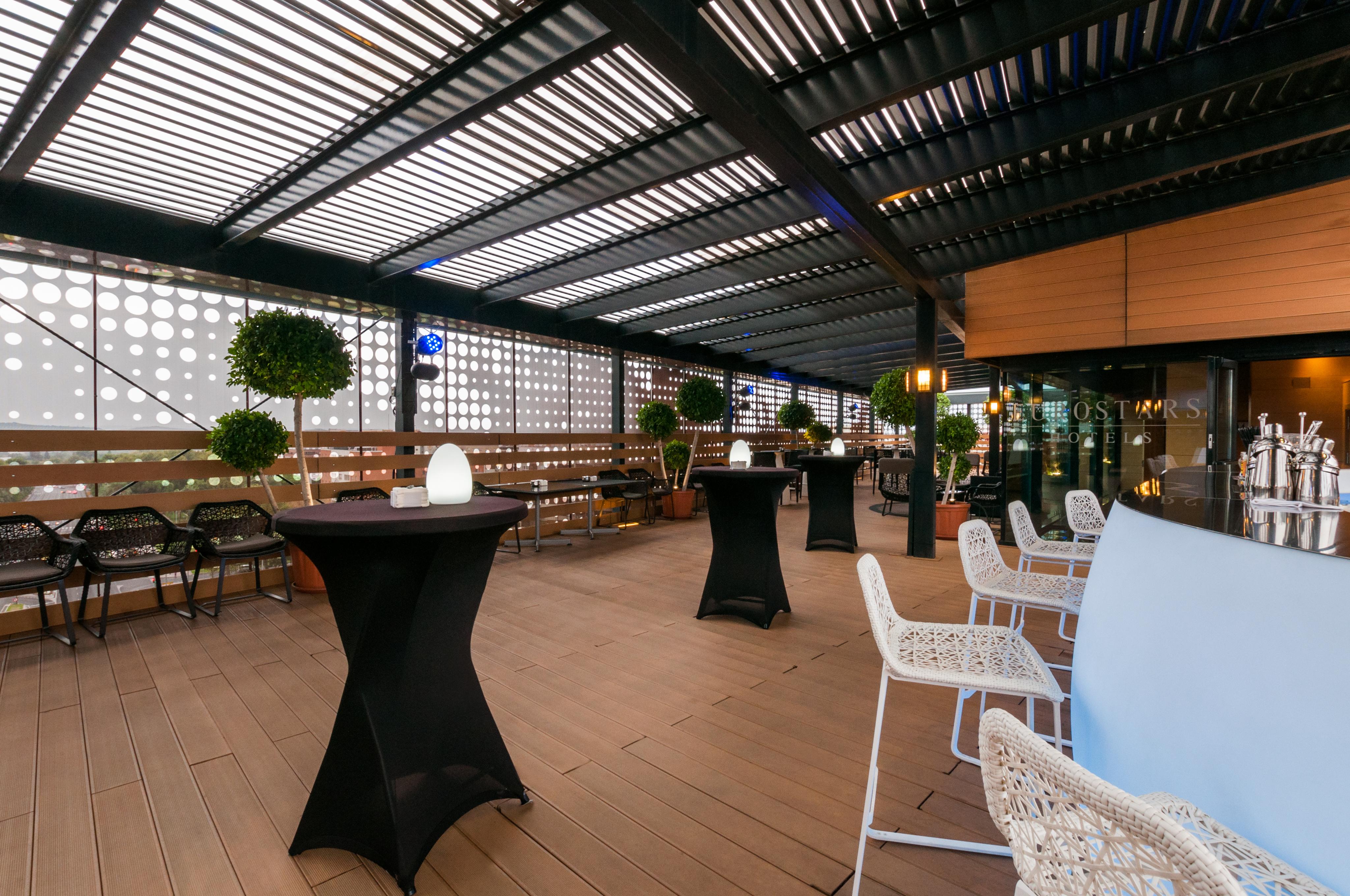Eurostars Palace Кордоба Екстериор снимка The photo shows a stylish outdoor seating area, likely part of a restaurant or lounge. It features a wooden floor and an overhead structure with horizontal slats, allowing for both shade and light. There are several tall, black cocktail tables and wh