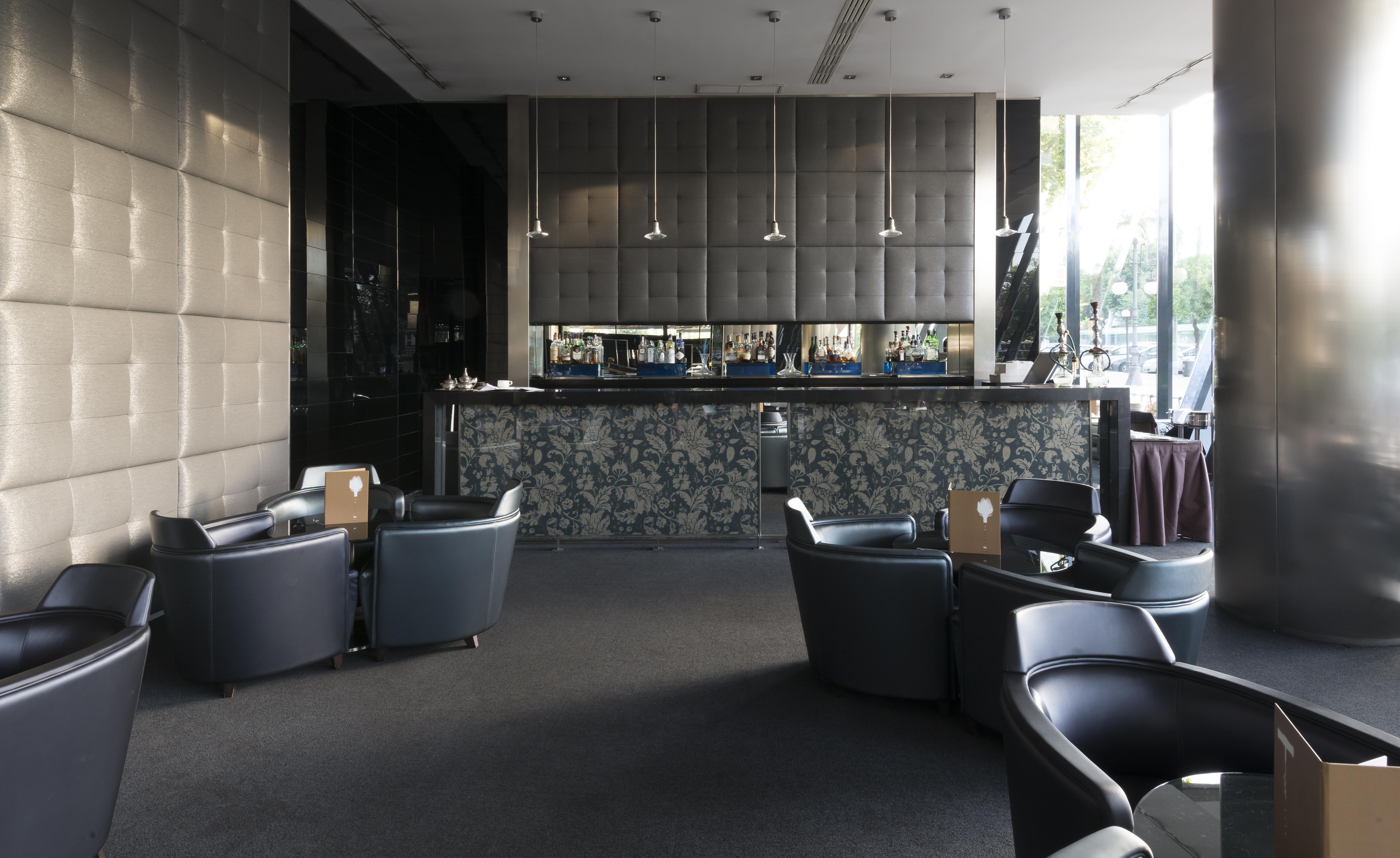 Eurostars Palace Кордоба Екстериор снимка The photo shows a modern bar or lounge area. In the foreground, there are several comfortable black armchairs arranged around small tables. The background features a sleek bar counter with a dark, textured wall behind it. The bar is illuminated with 