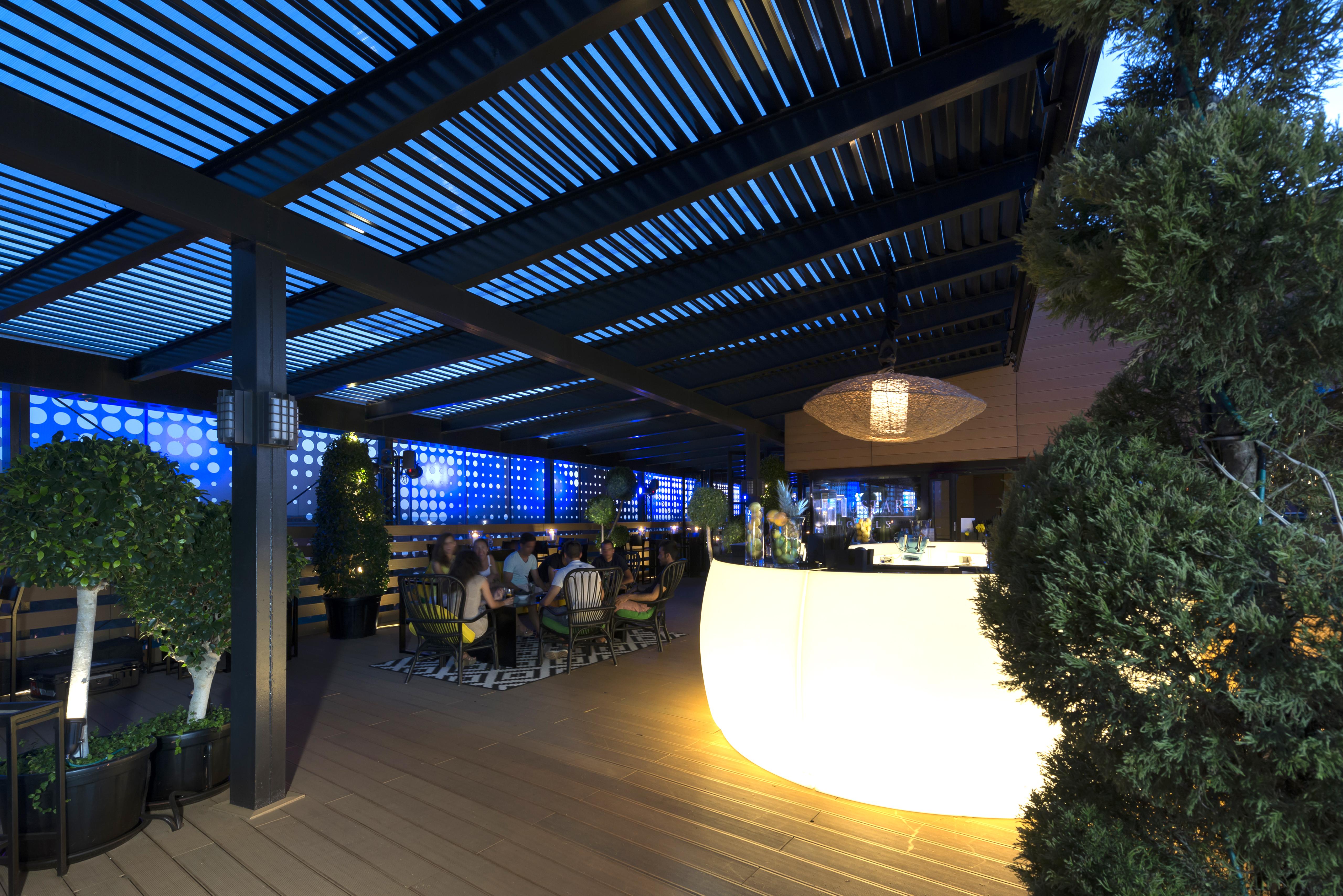 Eurostars Palace Кордоба Екстериор снимка The image shows a modern outdoor bar or restaurant area. The space features a sleek, illuminated bar that stands out with its curved shape. Patrons can be seen seated around tables, enjoying the ambiance. The overhead structure consists of slatted pa