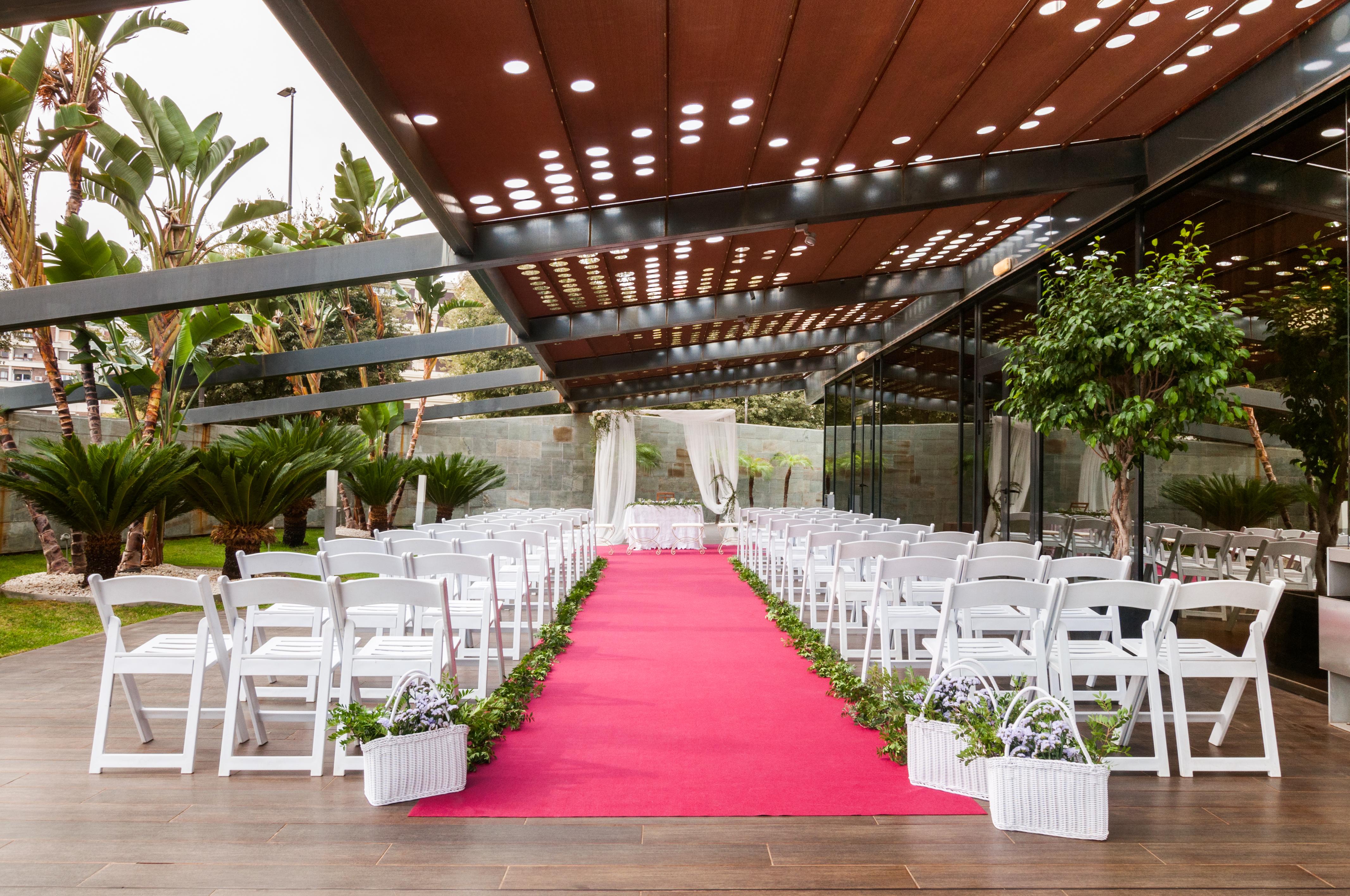 Eurostars Palace Кордоба Екстериор снимка The photo shows a wedding setup in an outdoor venue. There are white chairs arranged in rows on either side of a vibrant pink carpet leading to a floral arch or backdrop at the front. The area is partially shaded by a modern structure with a metal fr