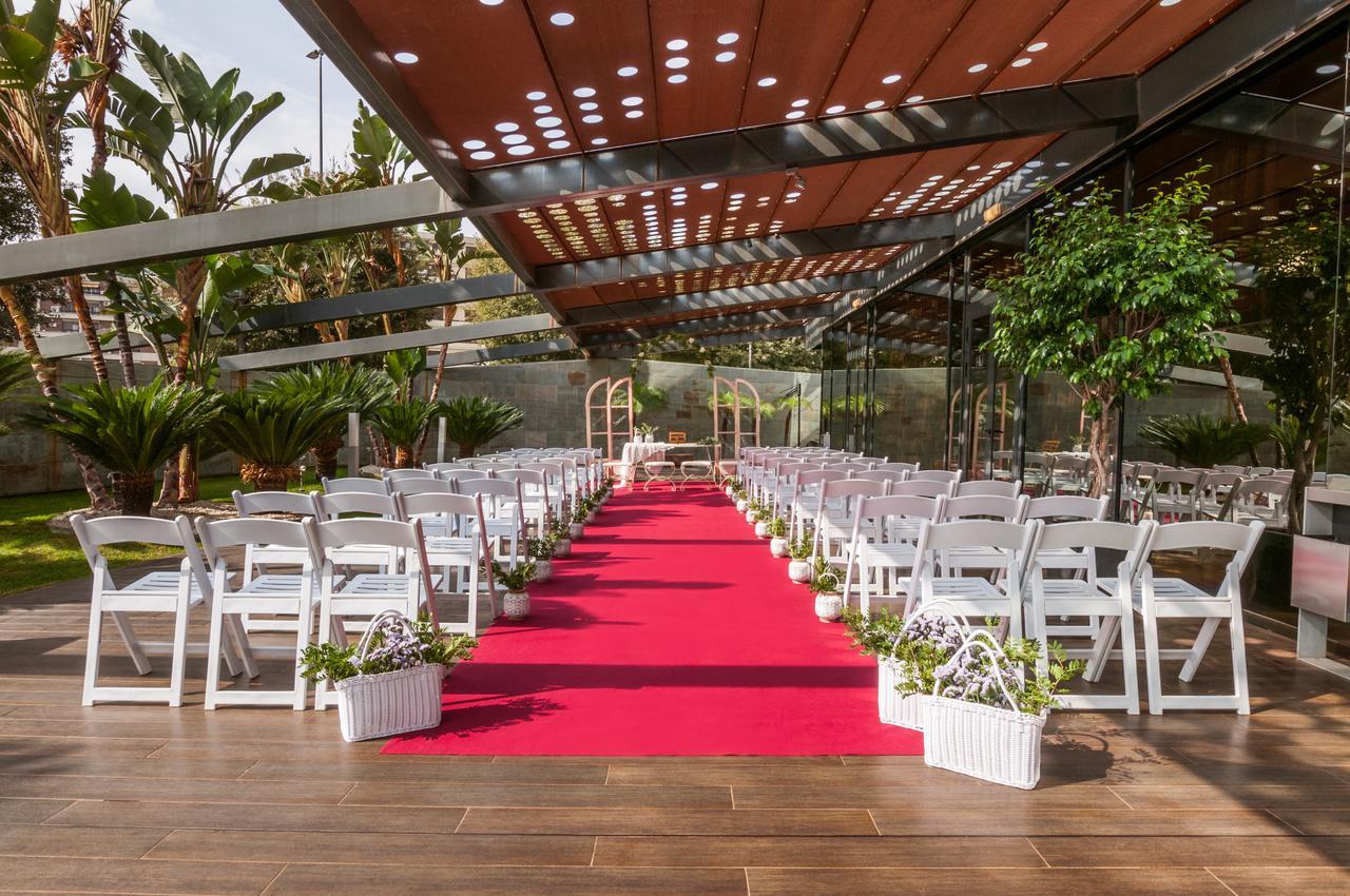 Eurostars Palace Кордоба Екстериор снимка The image shows an outdoor wedding setup. There are white chairs arranged neatly on either side of a vibrant red carpet that leads to a framed archway. The area is covered by a glass and metal structure, allowing plenty of natural light, while tropic